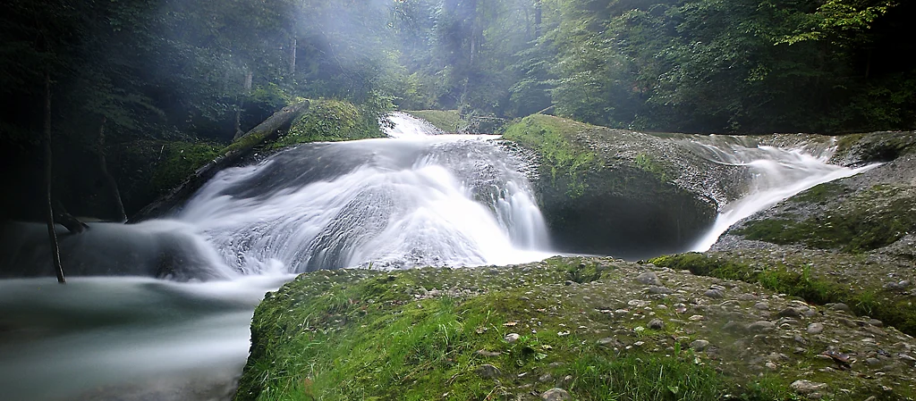 Wasserfall im Eistobel zwischen moosbewachsenen Felsen und grünem Wald